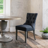 A charcoal grey padded dining chair with a button back and stud detail, standing on a wooden floor against a concrete background.