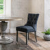A charcoal grey padded dining chair with a button back and stud detail, standing on a wooden floor against a concrete background.