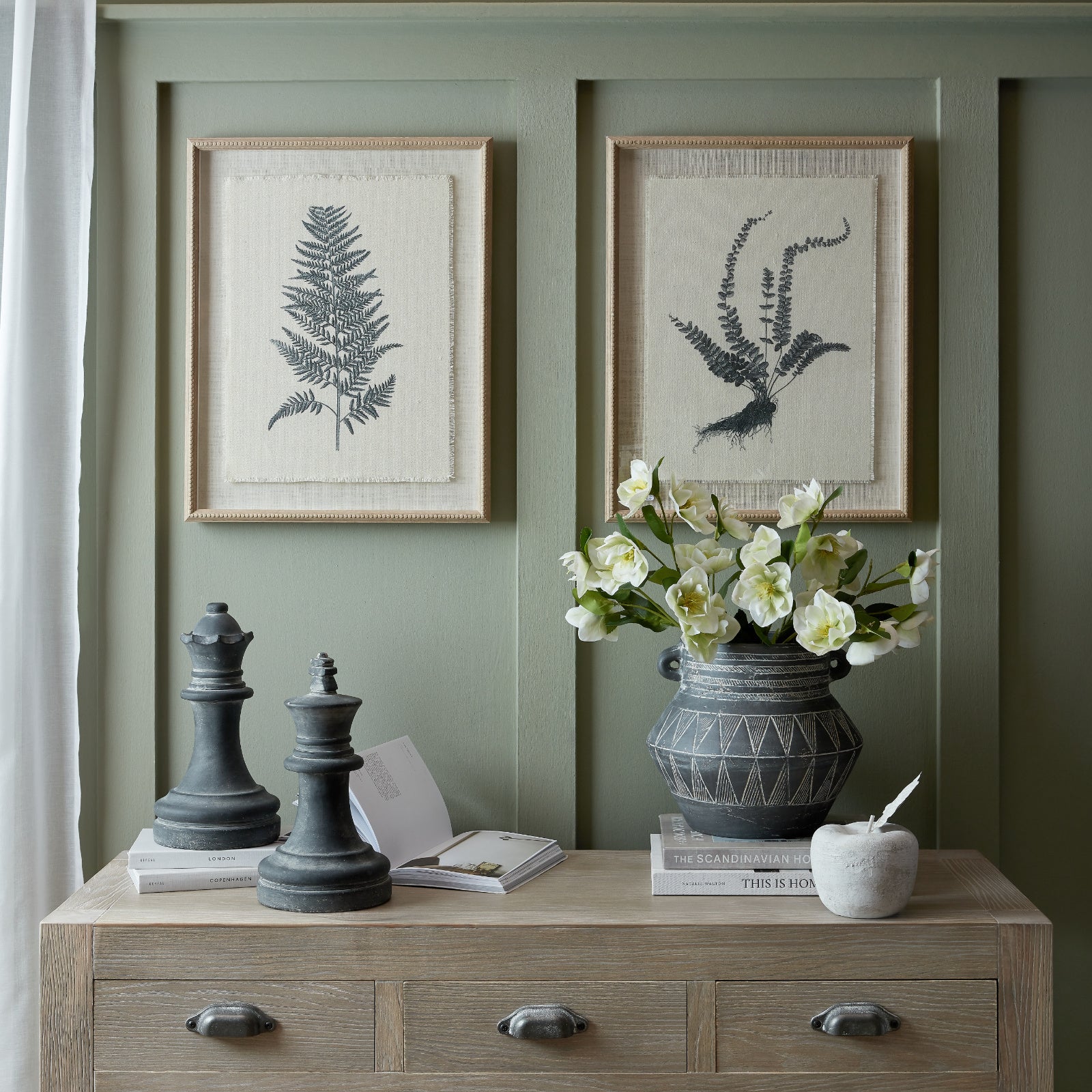 Decorative setup with framed botanical prints, a vase with flowers, and decorative items on a wooden cabinet against a green wall.