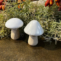 Two white ceramic mushrooms on a textured surface with greenery and red berries in the background.