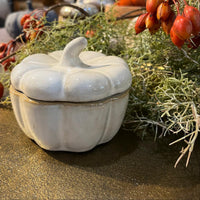 White ceramic pumpkin-shaped container with lid on a decorative surface with greenery and red berries.