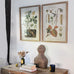 Two framed botanical prints on a wall above a wooden table with decorative items.